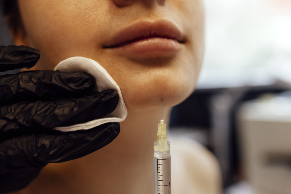 Close-up of a person receiving a cosmetic injection near the chin while a gloved provider holds a cotton pad against the skin.